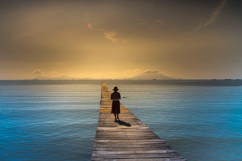 A solitary woman walks along a wooden pier at sunrise with calm ocean waters surrounding her.