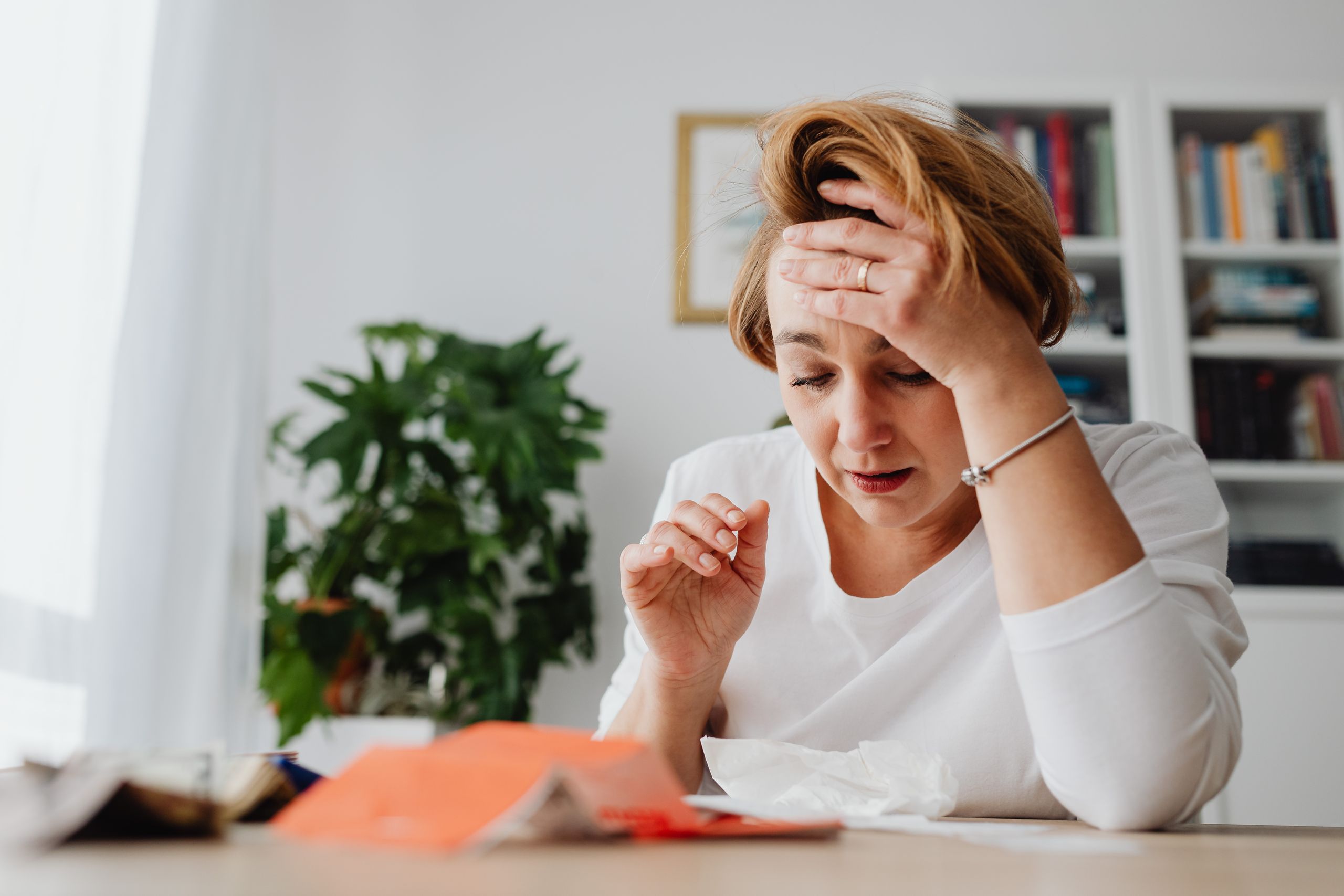 A stressed woman in a white shirt holds her head at a desk cluttered with papers, indoors.