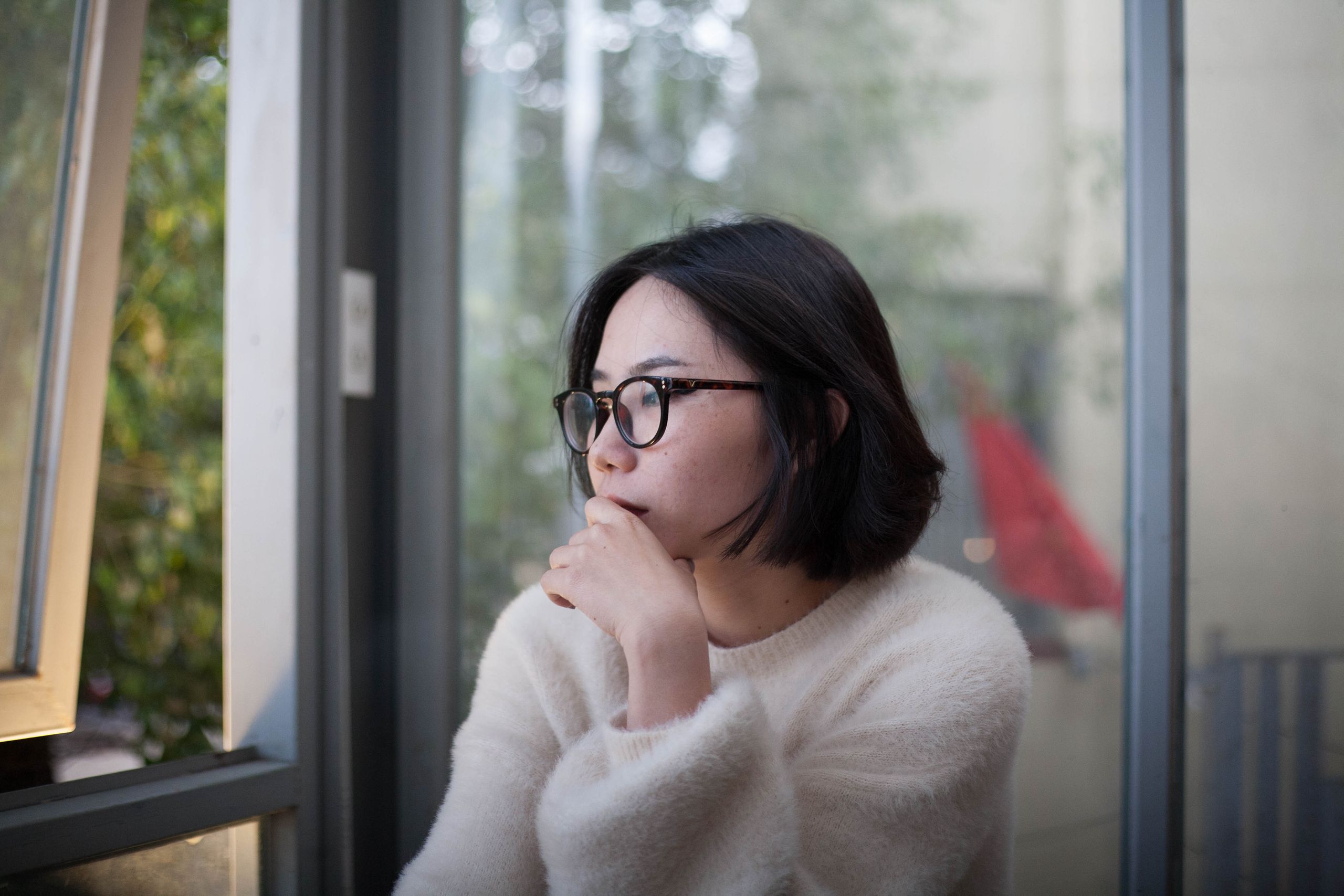 Asian woman reflecting in cozy sweater gazing thoughtfully out a window, hand on chin.