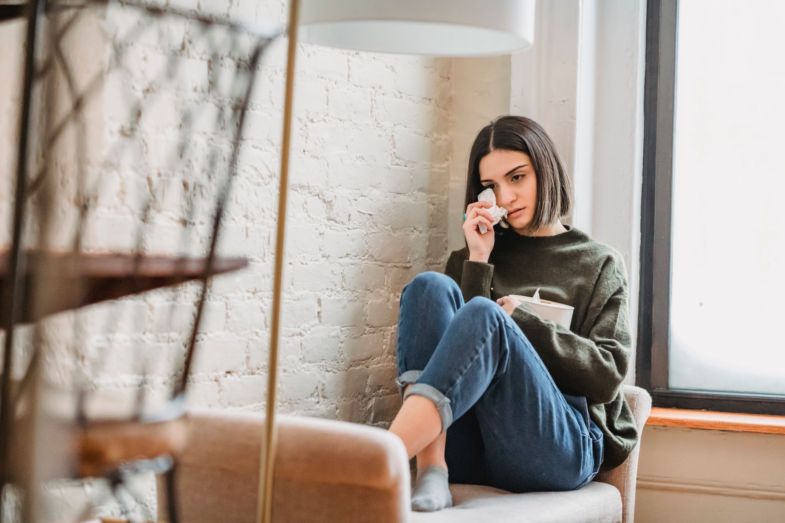 woman feeling hurt Full body of pensive young ethnic lady in casual clothes wiping tears with tissue while sitting on couch near window at home
